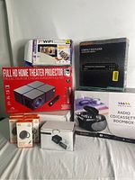 Overview photo showing all items in the bundle: home theater projector, digital photo frame, DVD player, radio boombox, Bluetooth speaker, and wireless chargers.
