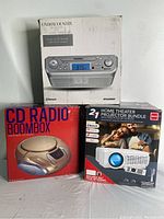 Three boxed items: Sylvania undercounter Bluetooth CD clock radio; CD radio boombox; RCA 2-in-1 home theater projector bundle with fold-up screen