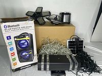 Photo showing Sylvania branded portable Bluetooth speaker in box, multiple LED solar lights, string lights, and a small solar panel all arranged on a table.