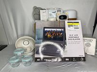 Photo shows boxed portable DVD player with 13.3 inch screen, a white small desk fan and four small containers with lids