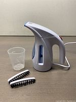 Front angle of white Hilife handheld garment steamer with measuring cup and two brush attachments on brown surface