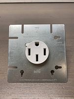 Square metal mounting plate with white 3-prong electrical outlet for range replacement part.
