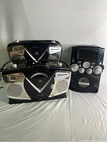 Two Sylvania retro style portable CD boomboxes and one Sylvania SKAR128 karaoke machine placed on a white surface, showing front view with controls and displays visible.