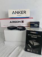 Boxes and devices including Anker SoundCore Bluetooth speaker box, Argon Helix gaming mouse pad box, Blackmagic SDI to HDMI micro converter box, Sabrent card reader box stacked on white packages, with converter and cables present.