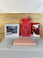 Front and side view of the pink power bank with foldable plug, the pink iPhone case, the Lexar 128GB microSD card in packaging, and the boxed pink digital camera on wooden table.