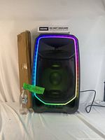 Front view of the ION PA Ultimate Freedom speaker with multicolor edge-glow lights illuminated and size reference with a bottle and a boxed speaker stand.
