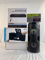 Photo showing stacked boxed units of HDMI upconvert DVD player, CD micro system with Bluetooth, and dual alarm clock radio, next to the Proscan Bluetooth speaker with RGB lights.