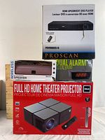 Boxes of RCA Full HD Home Theatre Projector, Wireless Bluetooth Speaker, Dual Alarm Clock with Radio, and HDMI Upconvert DVD Player stacked together.