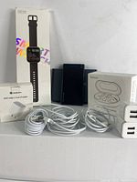 Photo showing the boxed smartwatch, wireless earbuds box, portable power bank, USB wall chargers, USB-C fast charger, and coiled USB to Lightning cables on a white surface.