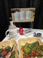 Group shot showing boxed canister set, hot chocolate pitcher with whisk, red tealight lamp, wine stopper gift box, two vegetable baskets