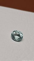 Oval natural blue aquamarine gemstone lying on a light textured surface, showing clarity and color from an angle.