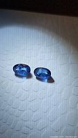 Pair of natural oval cut blue Ceylon sapphires placed on textured white surface under direct light, showing cut and color.