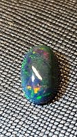 Oval black opal cabochon resting on textured surface showcasing multiple color flashes including green, red, and yellow.
