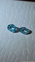 Two oval cut lagoon blue gemstones placed side by side on a textured white surface under light revealing facets and clarity.