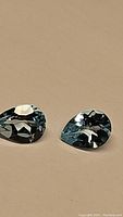 Close-up photo of two pear-shaped, faceted lagoon blue topaz gemstones placed on a beige surface, showing good clarity and cut.