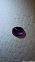 Close-up image of the oval cabochon cut purple ametrine gemstone on textured white surface, showing glossy finish and smooth contours.
