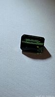 Close-up image of rectangular emerald-cut deep green chrome tourmaline gemstone placed on a white fabric surface showing its size, cut, and color.