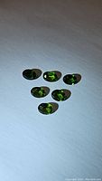 Six small oval faceted green gemstones displayed on white background under natural light showing color and cut.