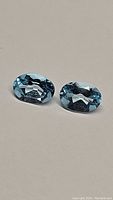 Pair of oval cut natural sky blue topaz gemstones side by side on a neutral background showing clarity and color.