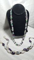 Two long semi-precious stone necklaces displayed on black and white backgrounds; stones vary in color including green quartz, amethyst chips, and quartz crystals.