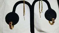 Front view of two pairs of gold plated earrings on black earring stand. Left side shows a pair of hoop earrings with red oval semi-precious stones embedded. Right side shows long oval drop earrings with diamond cut texture.