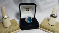 All three rings displayed on ring holders and in a black jewelry box showing the large blue stone ring.