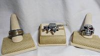 Wide shot of 3 stainless steel biker rings displayed on beige ring holders: Harley-Davidson logo ring, M16 machine gun ring, and skull ring with red crystal eye and cigar.