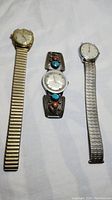 Three vintage men's watches laid flat: left gold-tone Bulova with expansion band, center watch with decorative sterling silver band with turquoise and red coral, right silver-tone Timex with expansion band.