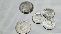 Five coins displayed on a white surface including one Morgan dollar, one Eisenhower dollar, and three Kennedy half dollars.