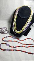 Multi-strand semi-precious stone chip necklaces displayed on black jewelry bust. Includes colorful strands of amethyst, peridot, citrine, and white pearls along with additional external necklaces with purple amethyst and red coral beads.