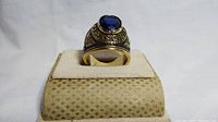 Front and top view of gold plated military ring with large blue stone on beige stand