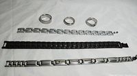 Photo showing 3 men's stainless steel bracelets and 3 rings arranged on a white surface in two rows; bracelets in bottom row and rings in top row.