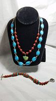 Full view of two necklaces on display stand and a bracelet laid out in front showing turquoise, red coral beads and silver details.