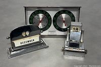 Three items shown: two vintage metal flip desktop calendars and one vintage Springfield desktop humidity and temperature gauge. All items have metal construction with some wear.
