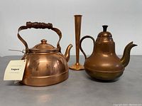 Front view of copper kettle with wooden handle, coffee pot with dark wrapped handle and dome lid, and tall narrow copper vase.