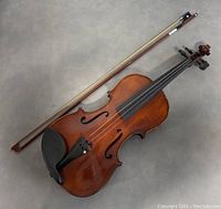 Violin displayed with bow on a gray floor background showing overall condition and details of the body and strings.