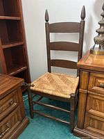 Single photo of antique wooden chair with woven wicker seat, ladder back, and finial tops on back posts, surrounded by wooden furniture pieces and a lamp.