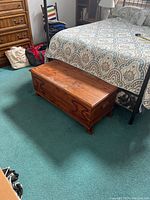 Photo of the wooden blanket box positioned at the foot of a bed on carpeted floor showing front and side views.