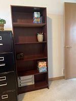 Front view of the wooden bookcase shelf with various small items on shelves, situated beside a black filing cabinet.