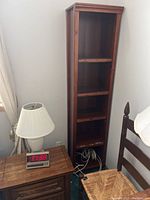 Tall narrow wooden bookcase beside a side table with lamp and clock, showing overall size and shape.