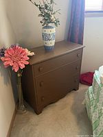 Full view of brown wooden chest of drawers positioned against wall next to a bed, with decorative vases and flower display on top.