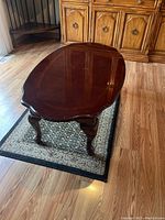 Wooden coffee table viewed from an angle showing polished oval top and cabriole legs, placed on a small patterned rug in a room with wooden flooring and a wooden cabinet in background.