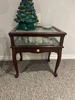 Full view of wooden end table with glass top, showing a green ceramic Christmas tree decoration on top and some items inside the table under the glass top.