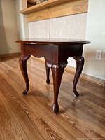 Side perspective showing the curved apron and Queen Anne style cabriole legs of the dark brown wooden round end table on a hardwood floor.