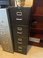 Front view of black metal file cabinet showing four drawers with label holders and handles, placed next to a beige file cabinet and wooden shelf.