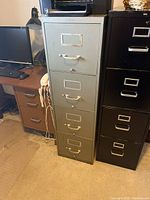 Front view photo showing the silver metal file cabinet with four drawers, each featuring a handle and label holder. Cabinet is situated between two other file cabinets and next to a desk with computer equipment.