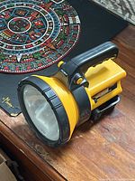 Photo of a yellow and black handheld flood flashlight with a large clear lens and ergonomic handle placed on a wooden surface.