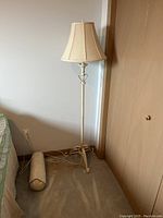 Floor lamp with beige fabric shade, slender metal post with decorative accent and tripod metal base. A beige cylindrical pillow is next to the base.