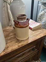 Photo showing a vintage ceramic jar sitting on a wooden dresser with brown and beige glaze and two small handles near the neck.