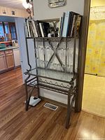 Front view of metal kitchen shelf located in kitchen setting, displaying top shelf with books and decorative items, two rows of hanging wine glass holders with glasses, and bottom rack for bottles or items.